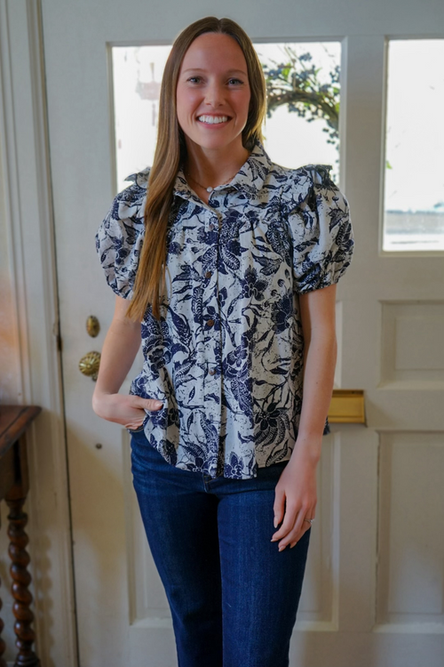 Model is wearing a navy and cream botanical print button down shirt with puff sleeves and ruffle detail.