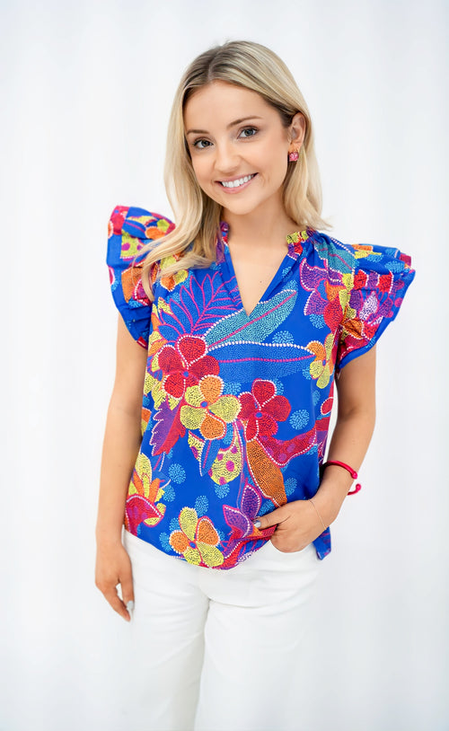 Model is wearing a cobalt blue, red and orange poplin top with a tropical print and ruffle sleeves.