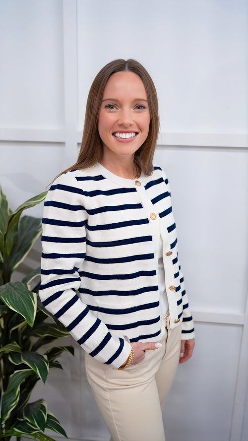 Model is wearing a preppy, long-sleeved blue and white sweater cardigan with gold buttons and patch pockets