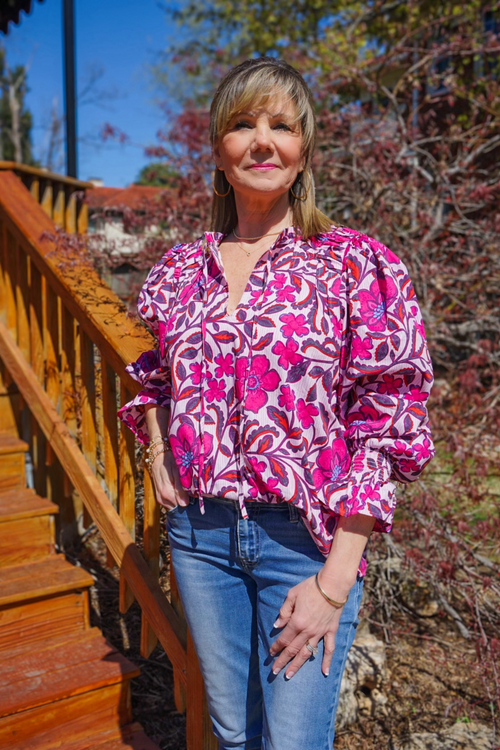 Model is wearing a pink floral poplin blouse with self tie at neck and long sleeves with smocking and ruffles at the wrists.
