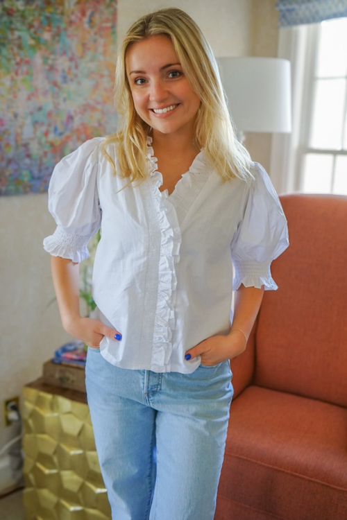 Model is wearing a white poplin blouse with ruffle detailing along the neck and along the placket. The blouse is fully lined and has puff sleeves with ruching and ruffle detail.