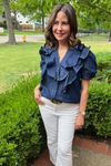 Model is wearing a dark denim button down blouse with layered ruffles along the bodice and puff sleeves. Flattering v-neck design.