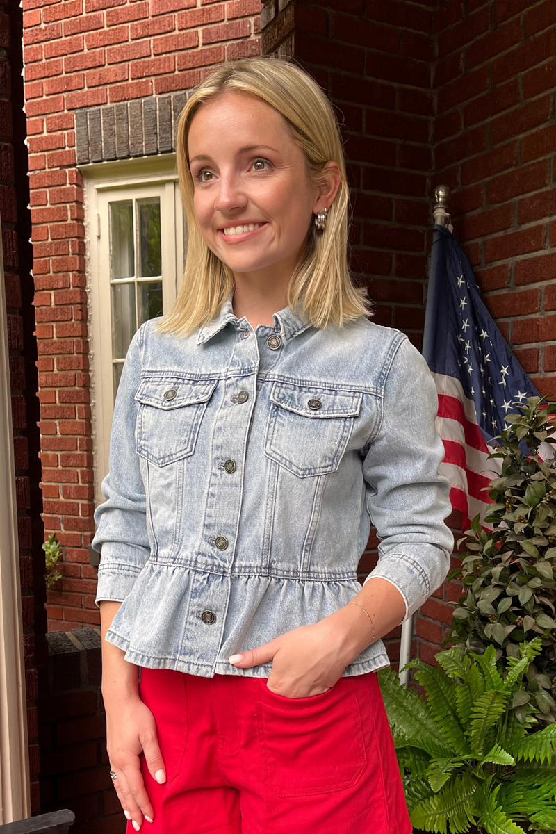 Model is wearing a light wash denim jean jacket with patch, flap button closure pockets and peplum at the waist. 