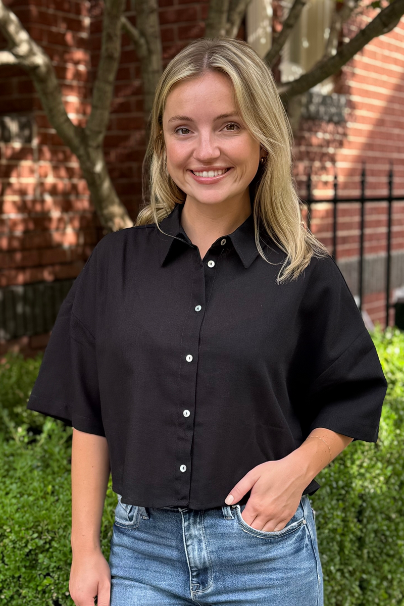 Model is wearing a black button down short sleeved top with boxy, cropped fit.