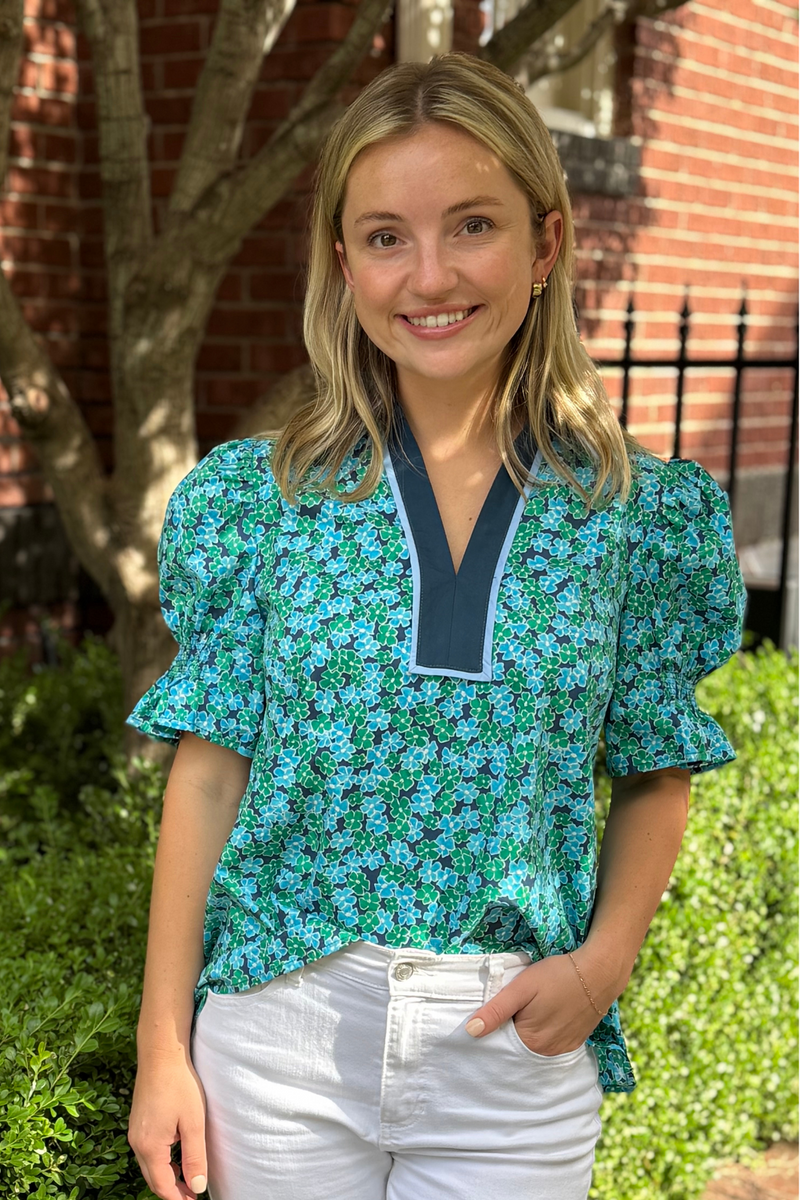 Model is wearing a blue and green ditsy print floral blouse with v-neck and puff sleeves.