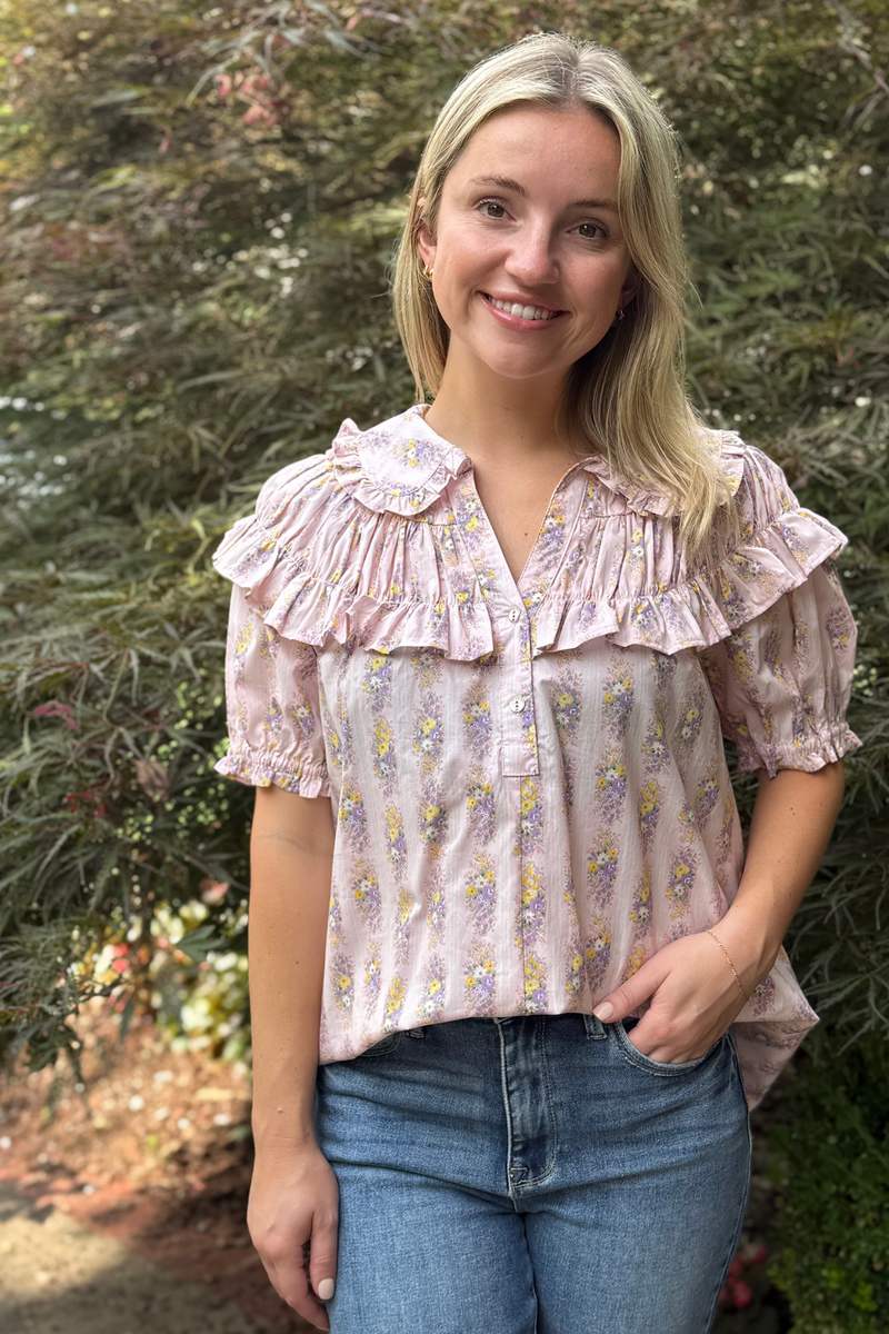 Model is wearing a light pink floral print top with pintucked and ruffle collar and short puff sleeves.
