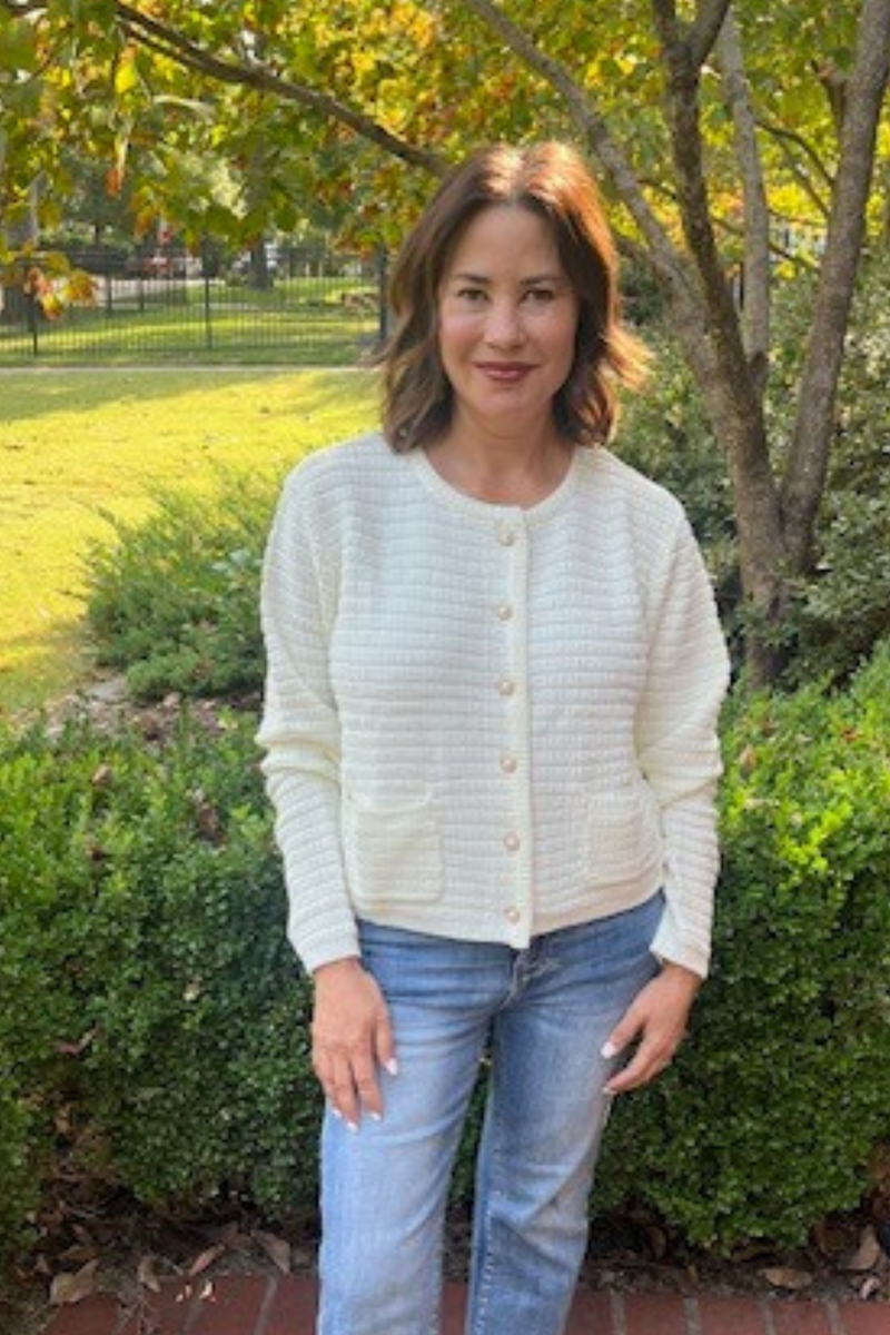 Model is wearing a cream colored cardigan with front patch pockets and pearl functional buttons. 