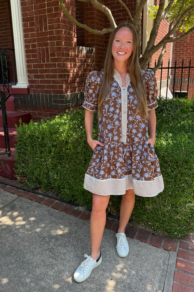 Model is wearing a chocolate, blue and cream floral dress with blue and cream ticking detail along the hem, sleeves, collar and placket.