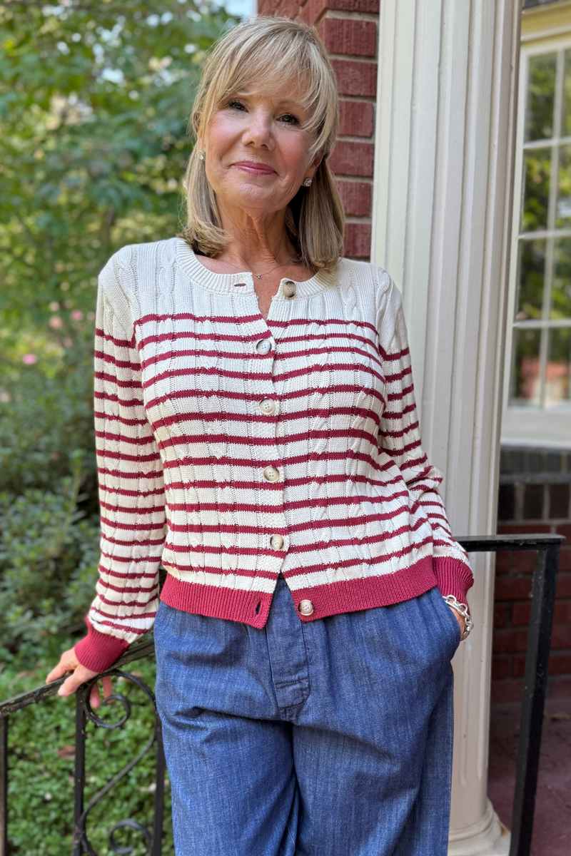 Model is wearing a lightweight cream cable knit cardigan with burgundy stripe detailing.