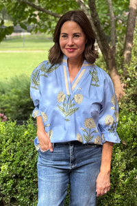 Model is wearing a periwinkle blue shirt with blue and yellow tape detail along the v-neckline and yellow and green botanical pattern throughout.