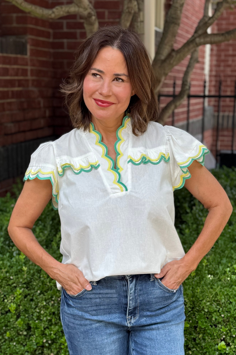 Model is wearing a white short sleeve cotton blouse with yellow and green scalloped piping along the sleeves, collar and bodice.