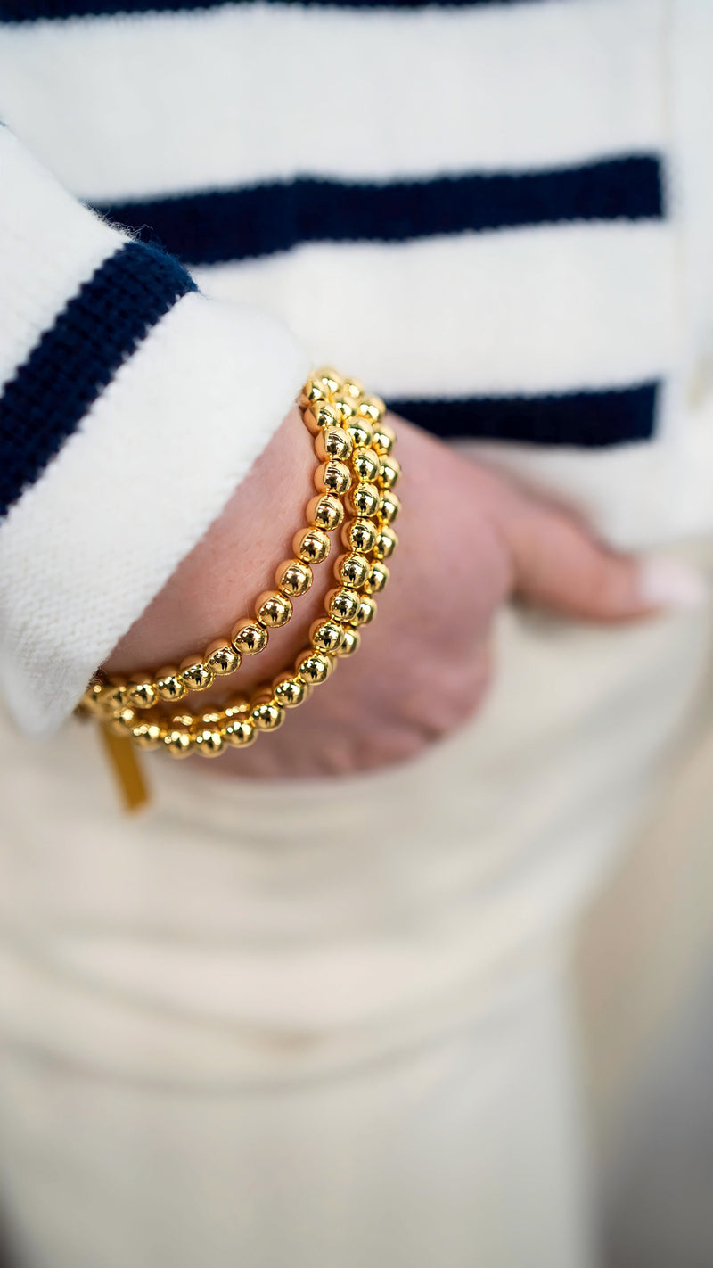 Model is wearing set of three gold beaded bracelets with stretch construction