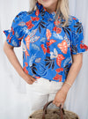 Model is wearing  blue, red and orange tropical print blouse with puff sleeves and ruffle collar.