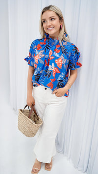 Model is wearing a blue tropical print popover blouse with ruffle collar and puff sleeves.