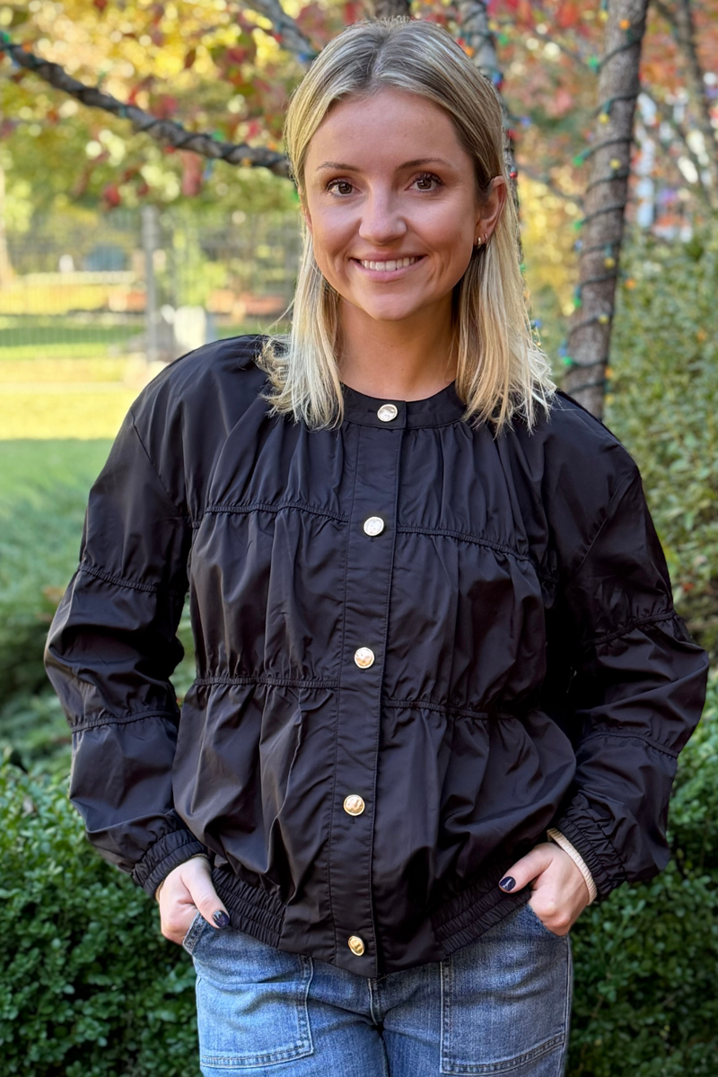 Model is wearing a black nylon jacket with stitch detailing with gold functional buttons down the front. Collarless design with elasticized banding at the waist and wrists.